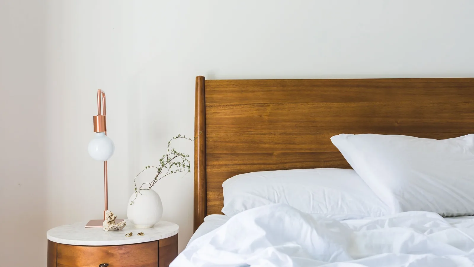 A wooden bed headboard with white bedding and a copper lamp on a bedside table.