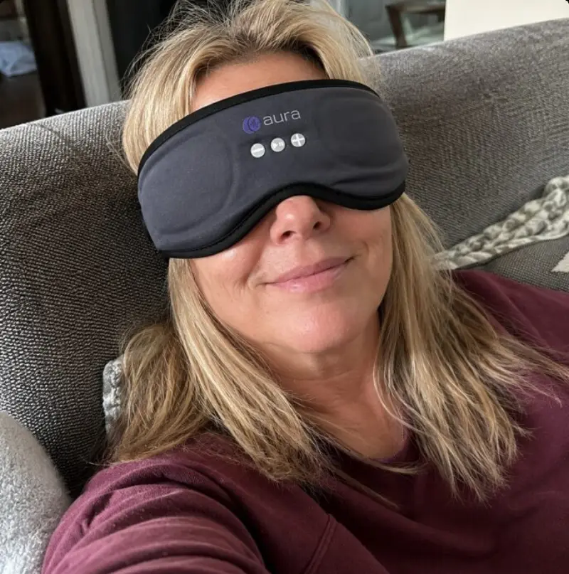 A woman in her fifties resting on a sofa wearing the grey AURA sleep mask with headphones.