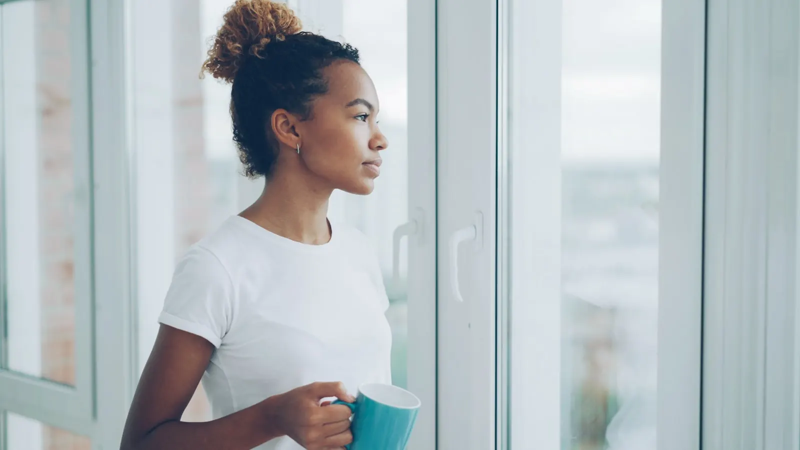A woman holding a warm mug, looking out a window in quiet morning light.