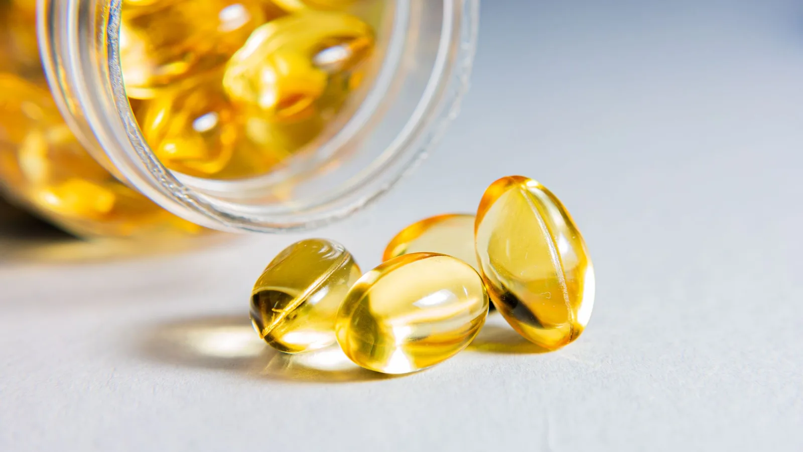 Amber supplement softgels spilling from a glass jar onto a pale surface.