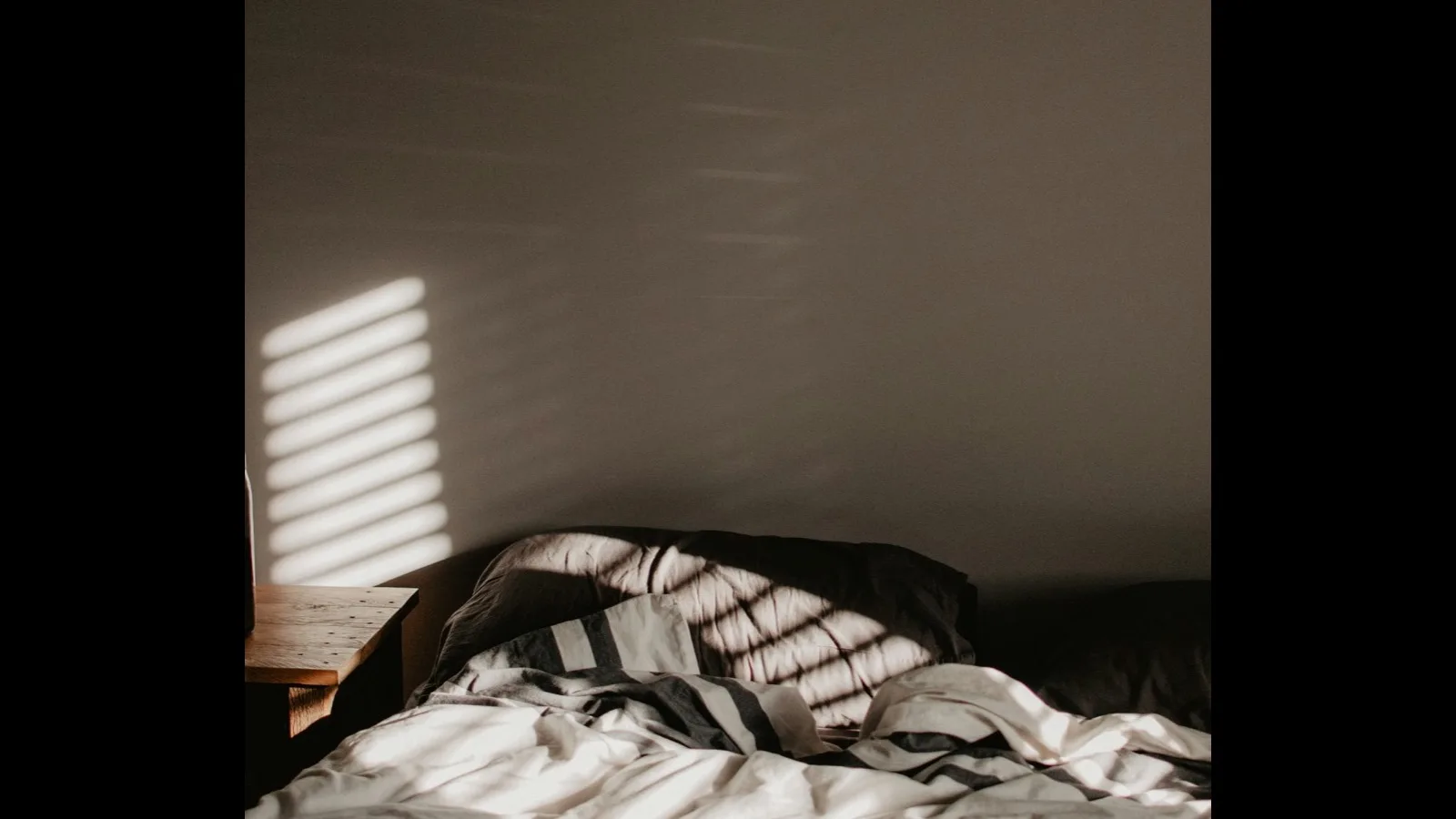 A rumpled, unmade bed in morning light coming through slatted blinds.