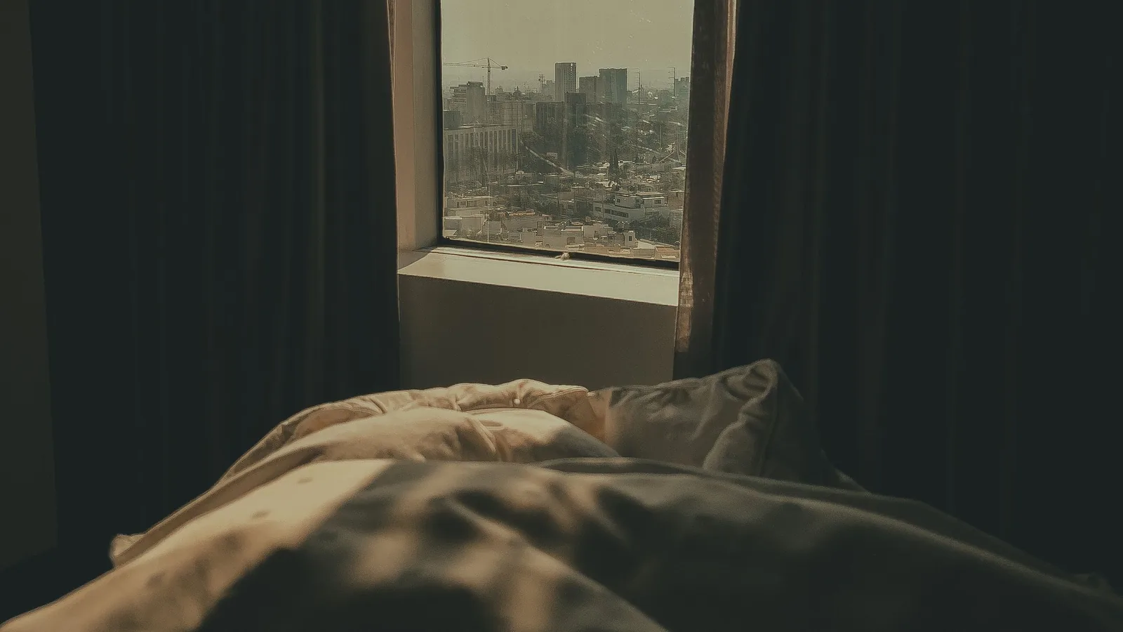 A dark bedroom with heavy curtains, a rumpled bed in the foreground and a city visible through the window.