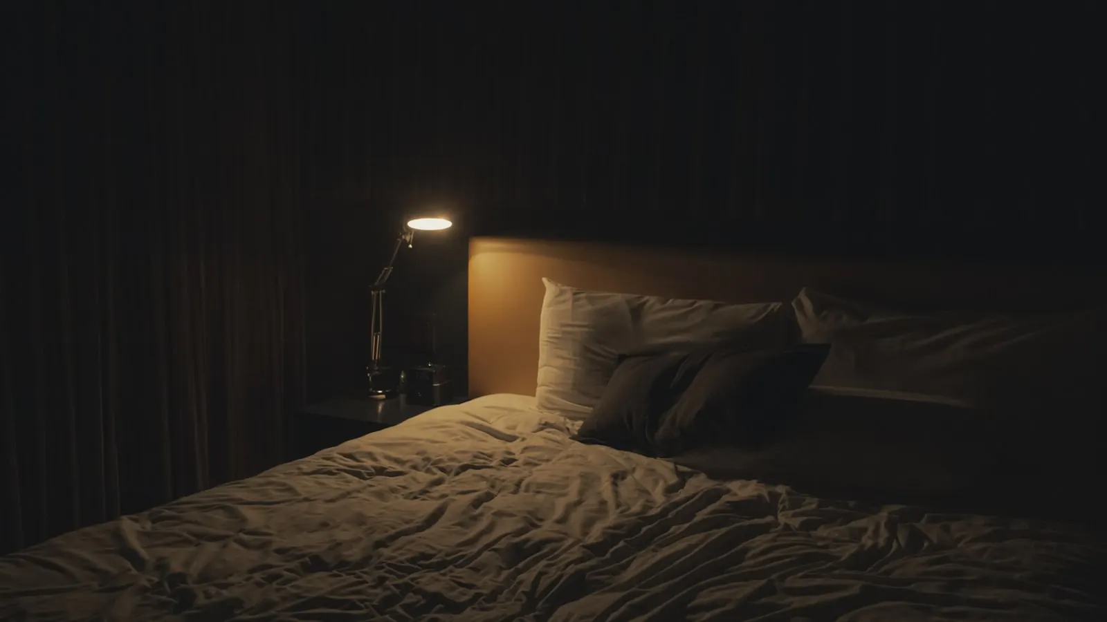 A warm lamp glowing beside a bed in an otherwise dark room, the kind of low light that's on at 3am.