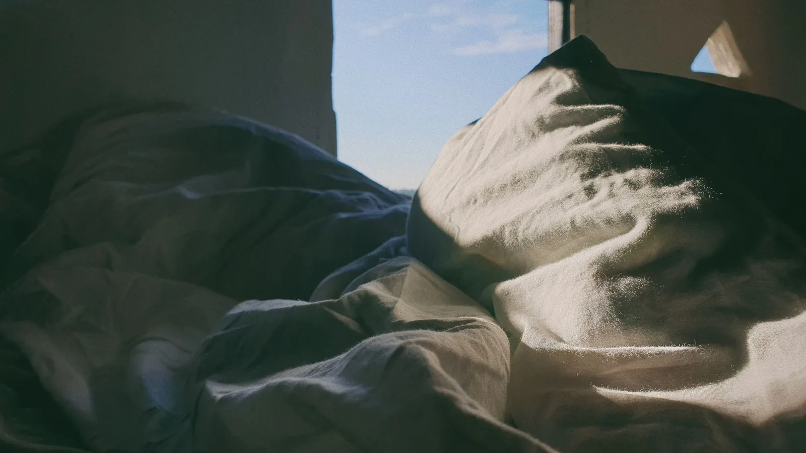 Crumpled bedding in dim light with a window open to a pale sky.