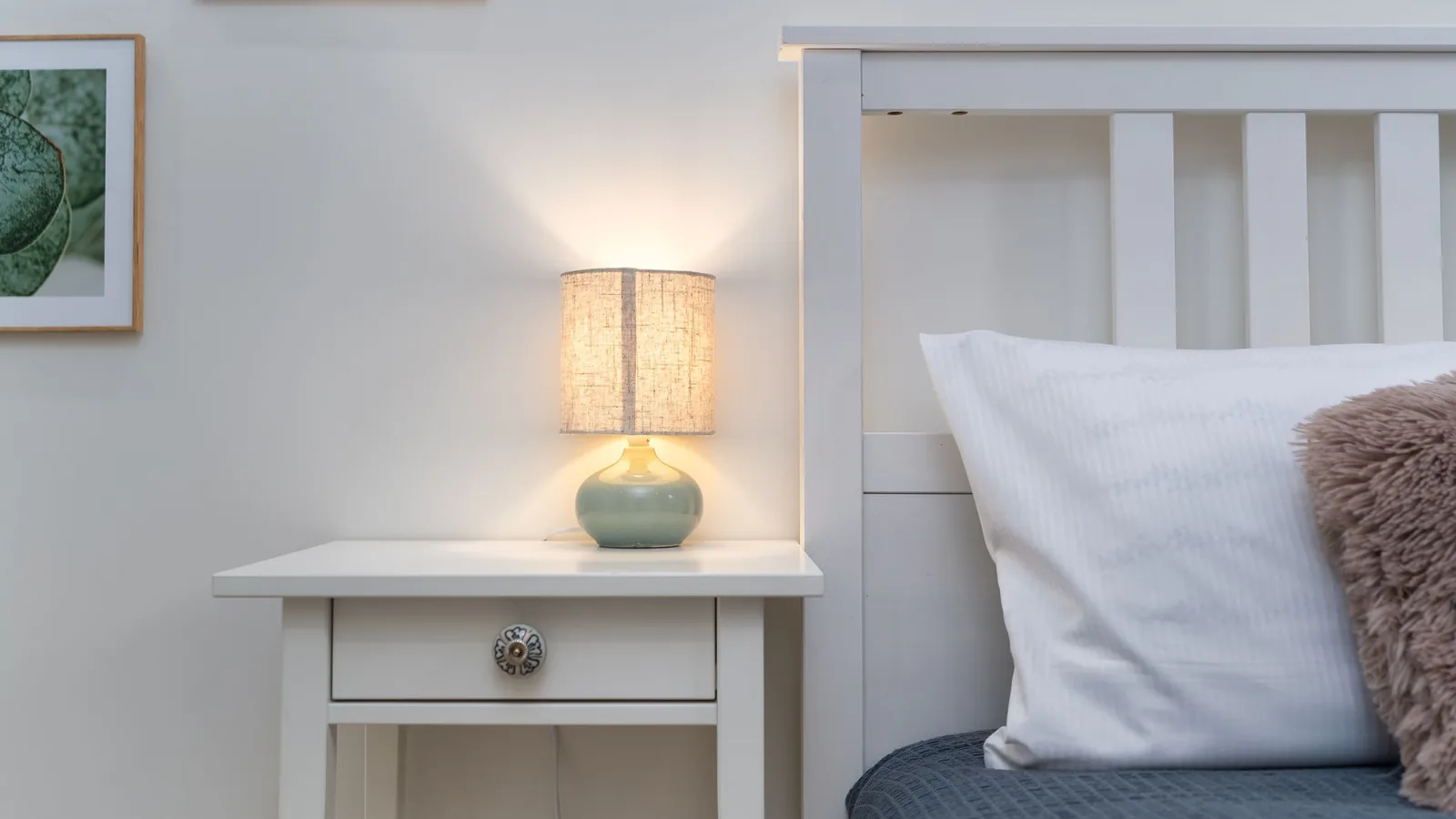 A lit bedside lamp on a small white nightstand beside a bed.